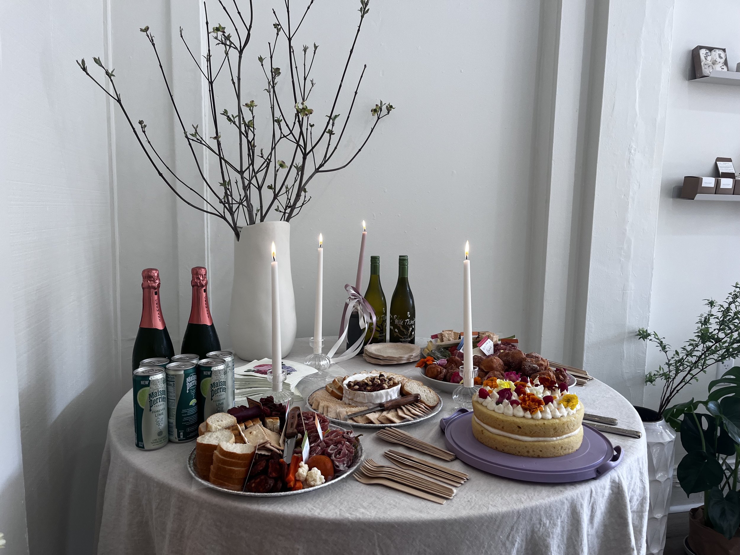 A table set for celebration with a cake, candles, various drinks, and snacks, decorated with a vase holding branches.