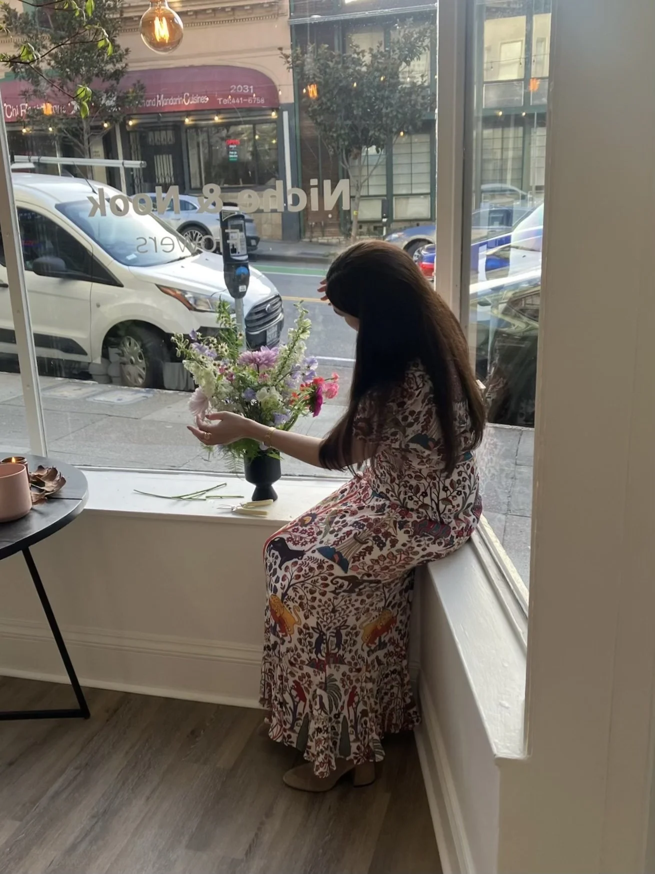 A woman sitting on a windowsill arranging a colorful bouquet of flowers inside a cafe.