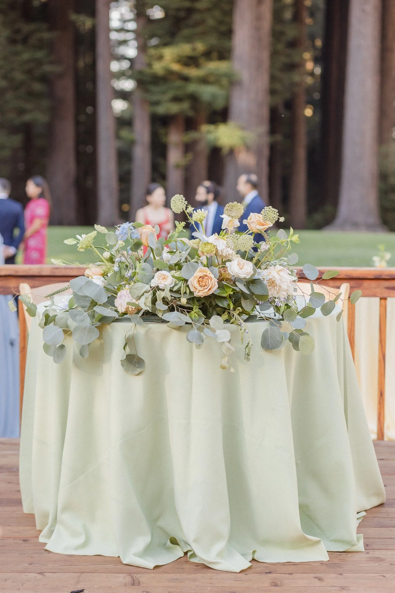 A flower arrangement on a table with light-colored cloth, set outdoors with tall trees and a group of people in the background.