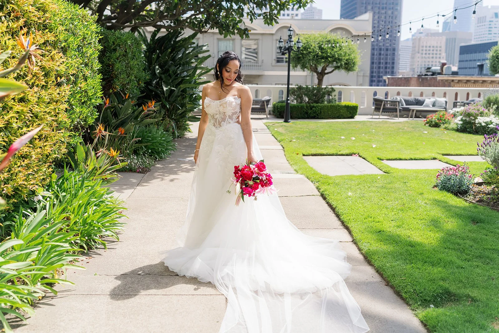 Bride in a white wedding dress holding pink flowers walking on a garden sidewalk with city buildings in the background.