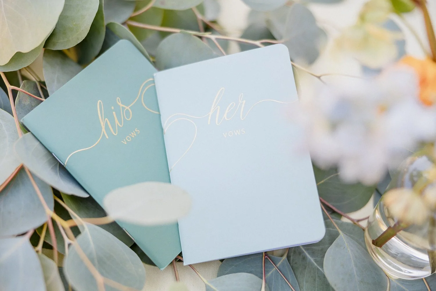 Two vow booklets, one labeled "her vows" and the other "his vows" in gold lettering, surrounded by eucalyptus leaves and placed on a light surface.