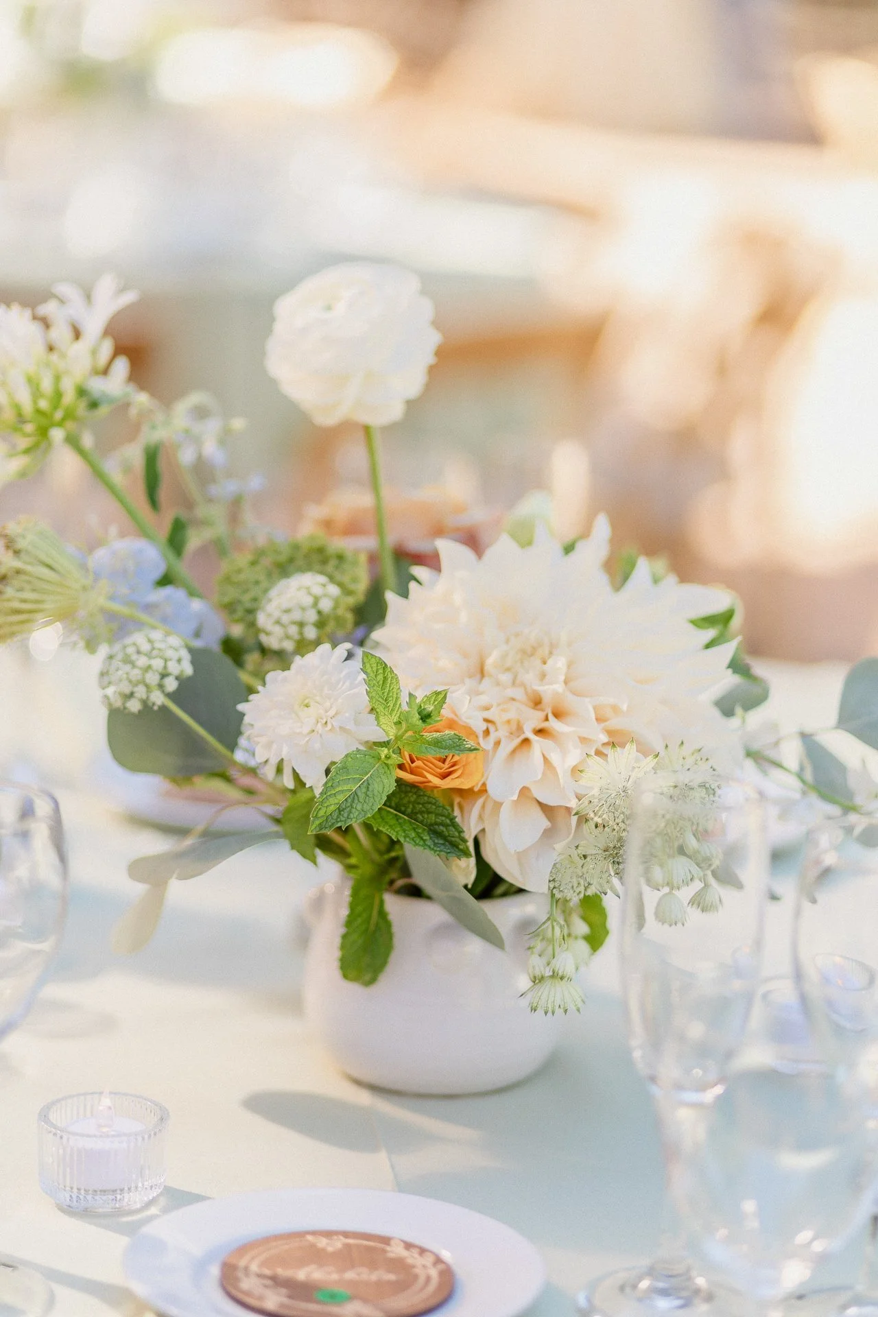 A floral centerpiece with white, cream, and peach flowers, green leaves, and eucalyptus on a table set for a celebration or wedding.