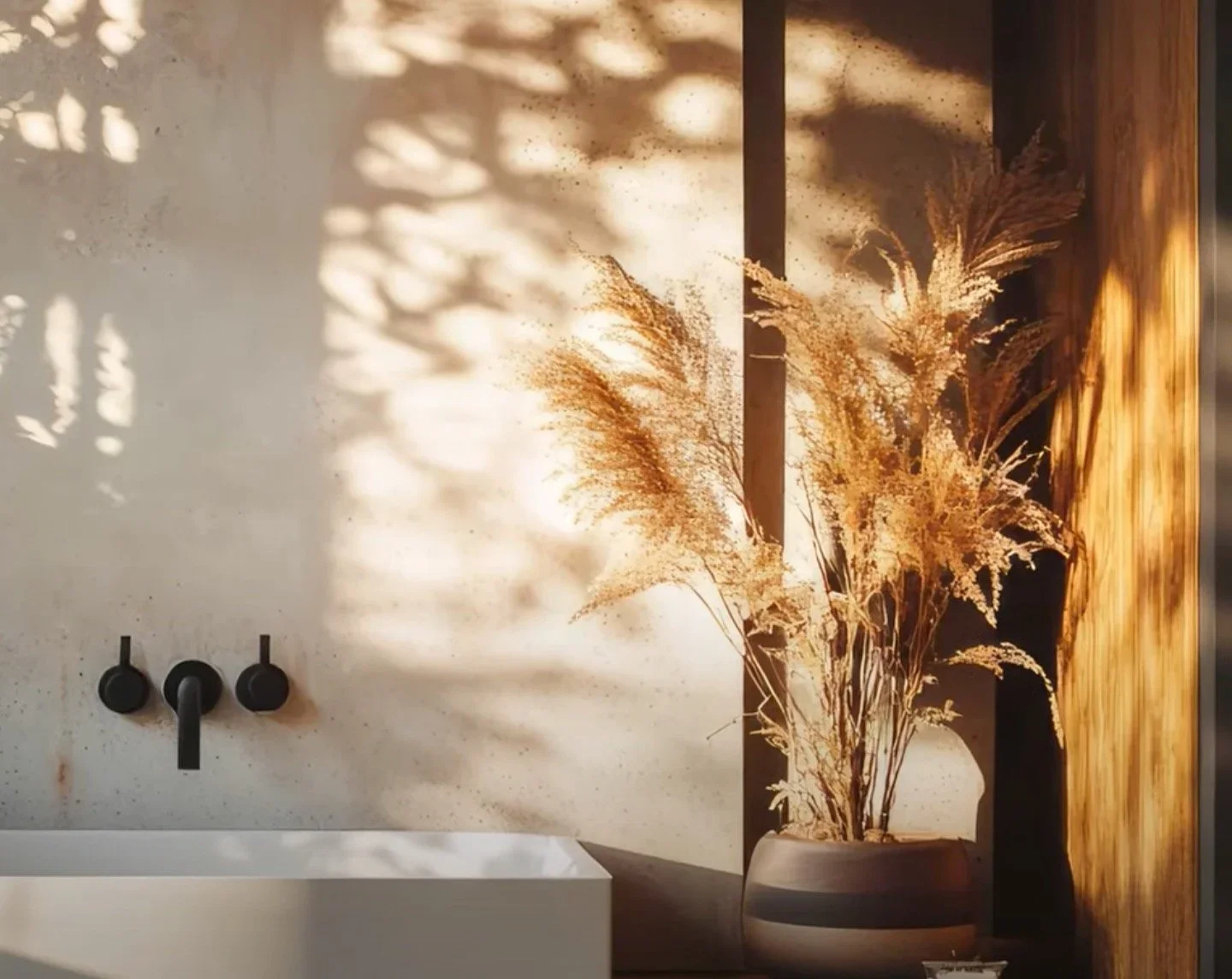 A minimalist bathroom with a white sink and a black faucet, decorated with a large beige vase of dried pampas grass. Shadows from a window create patterns on the wall.