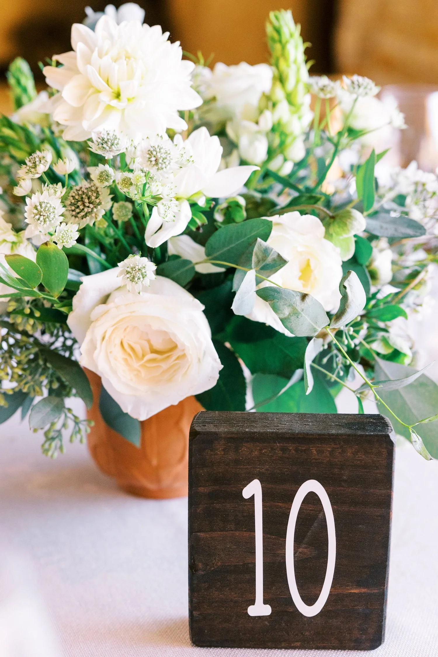 A floral centerpiece with white flowers and greenery in a brown vase, with a dark wood table number sign showing the number 10.