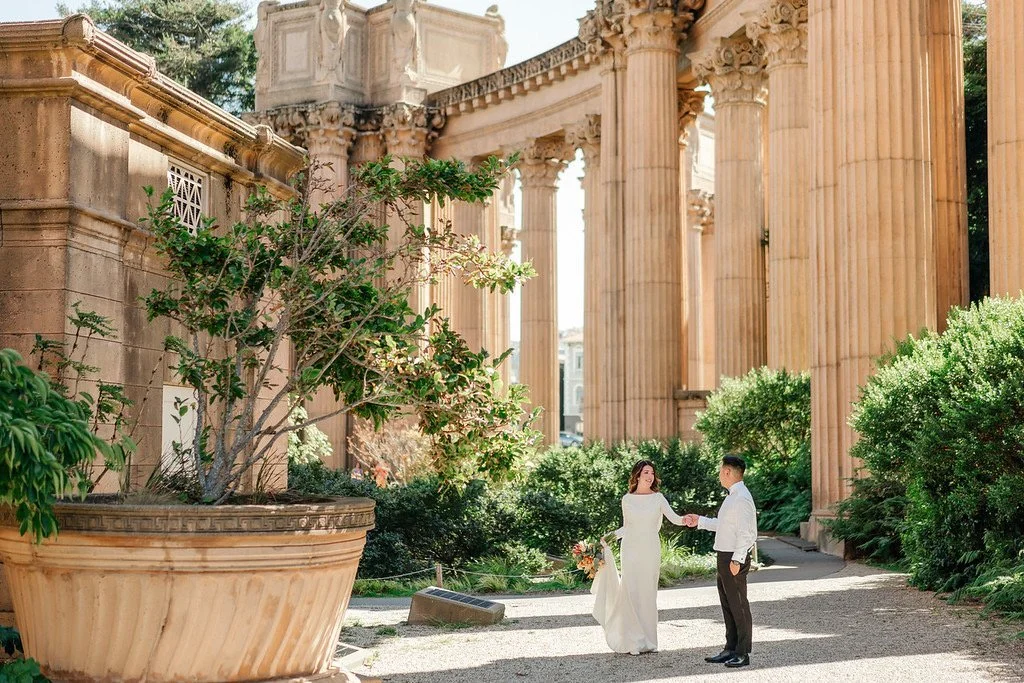 A bride and groom holding hands in front of ancient stone ruins with tall columns and lush greenery.