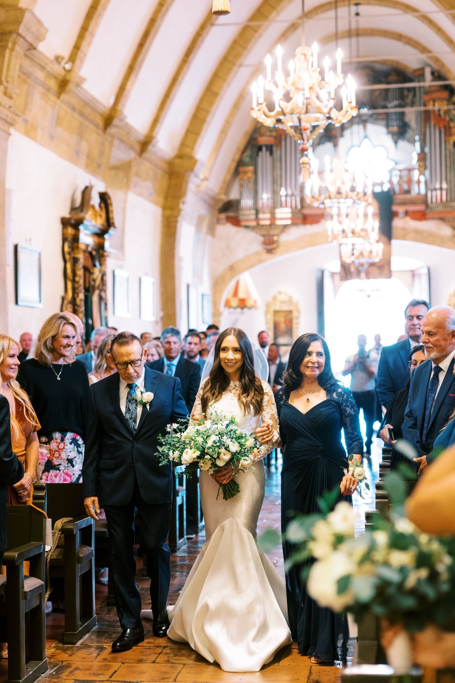A bride in a white wedding gown walks down the aisle in a church, accompanied by her parents. Guests are seated on either side, watching and smiling. The church is decorated with large chandeliers and wood accents.