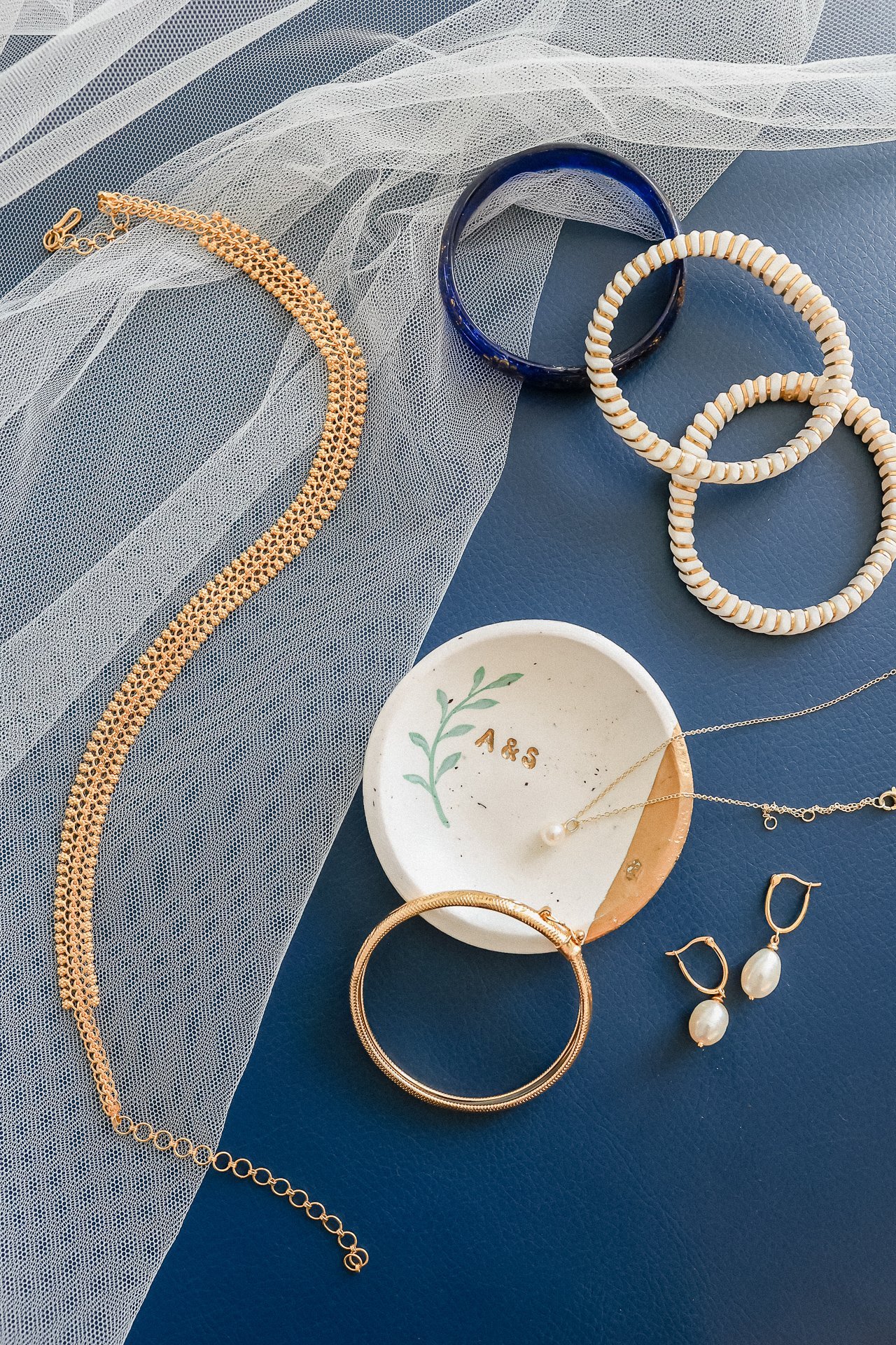 Various jewelry items including gold and pearl earrings, bangles, necklaces, and bracelets arranged on a navy blue surface with a piece of sheer white fabric and a small dish with the initials 'A&S' and a green leaf design.