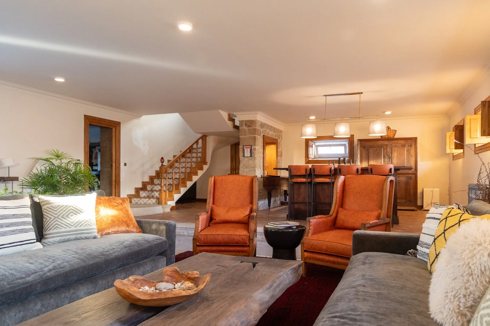 Spacious living room with gray and orange seating, wooden coffee table, and decorative pillows. In the background, a bar area with bar stools, a wooden cabinet, and a small window. A staircase with wooden banister leads upstairs, and a stone wall is visible near the staircase. Soft lighting and warm tones create a cozy atmosphere.