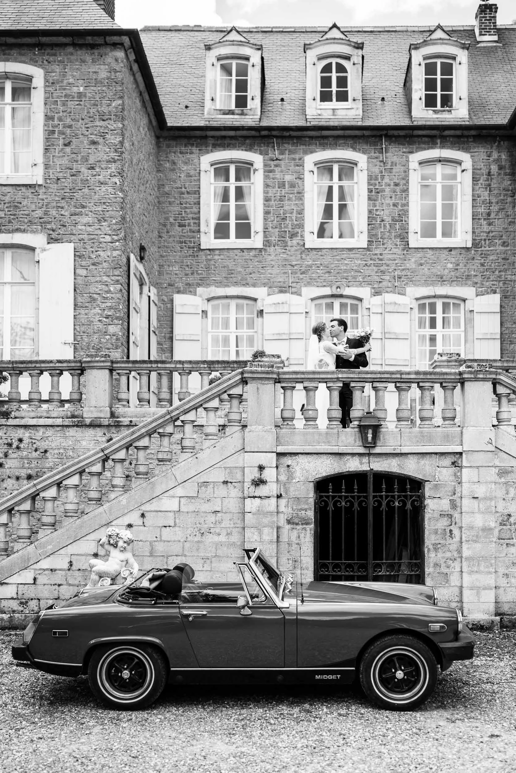 Photo en Noir et Blanc représentant un couple de marié qui s'embrasse devant le Chateau du Denacre dans le Boulonnais. Une voiture de collection les attend en contrebas