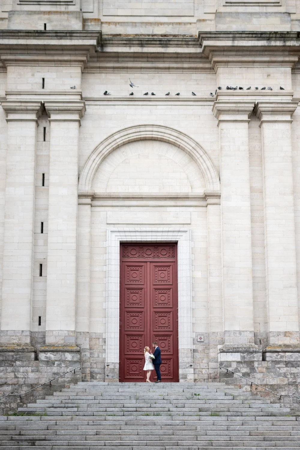 Photo d'un couple de mariés devant les portes rouge de la cathédrale Notre-Dame de l'Assomption et Saint Vaast d'Arras