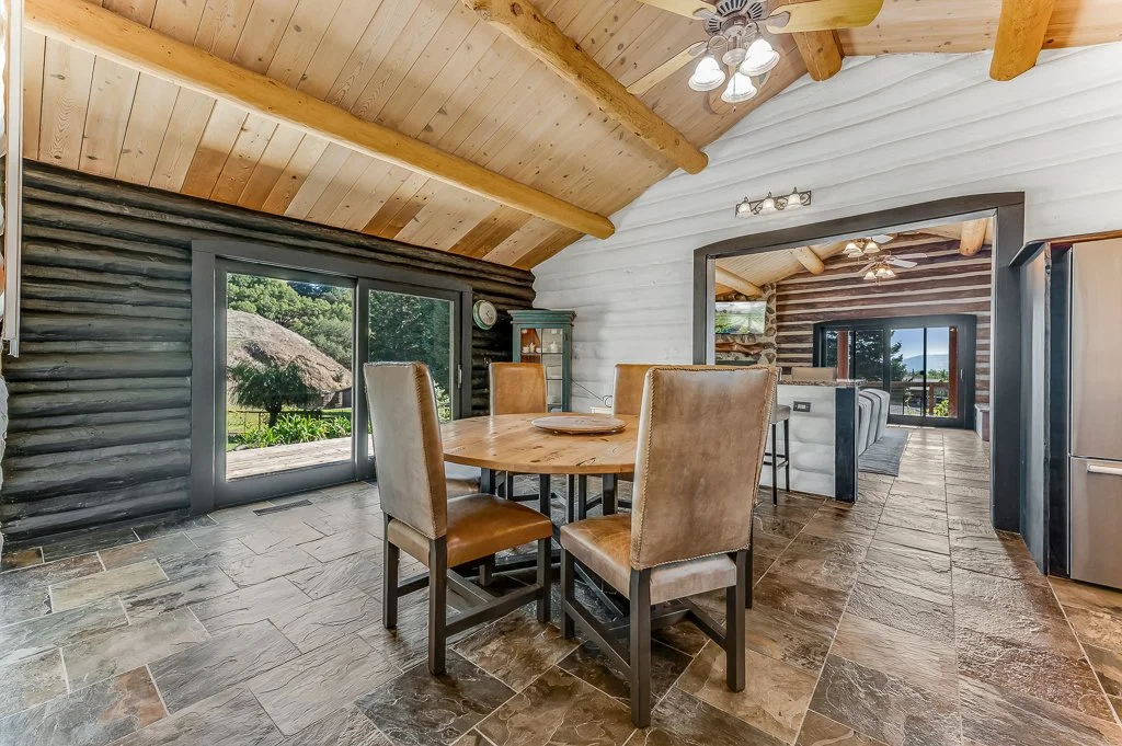 Interior of a rustic home with stone and wood walls, wooden ceiling with exposed beams, a round dining table with four chairs, sliding glass doors to greenery, and a glimpse of a living area with a TV and ceiling fans.