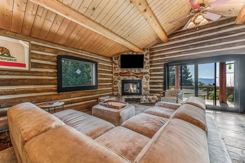 Living room with log cabin style interior, featuring a plush tan sectional sofa, stone fireplace with television above, wood ceiling with exposed beams, window with outdoor view, and chalet-style decor.