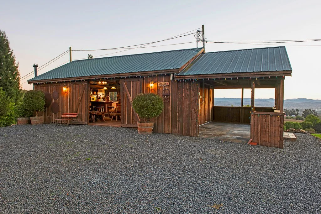 A rustic wooden house with a metal roof, outdoor lighting, and a gravel driveway overlooking scenic hills at sunset.