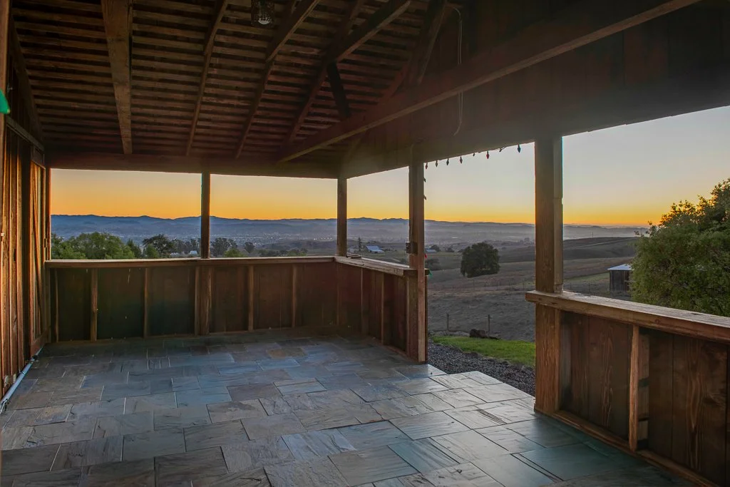 View from a wooden porch overlooking rolling hills during sunset with trees and distant mountains.