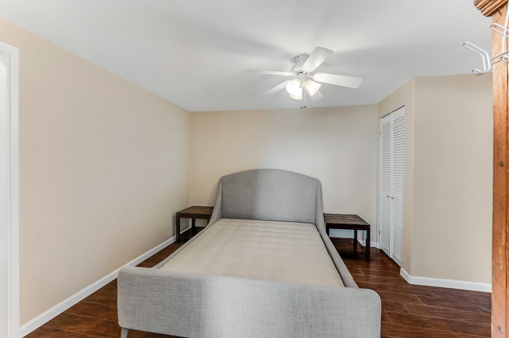 Simple bedroom with a gray upholstered bed, two wooden side tables, a ceiling fan with lights, beige painted walls, hardwood flooring, and a white closet door.