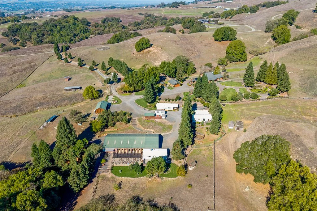 Aerial view of a rural estate with multiple houses, a barn, and trees spread across rolling hills with dry and green patches.