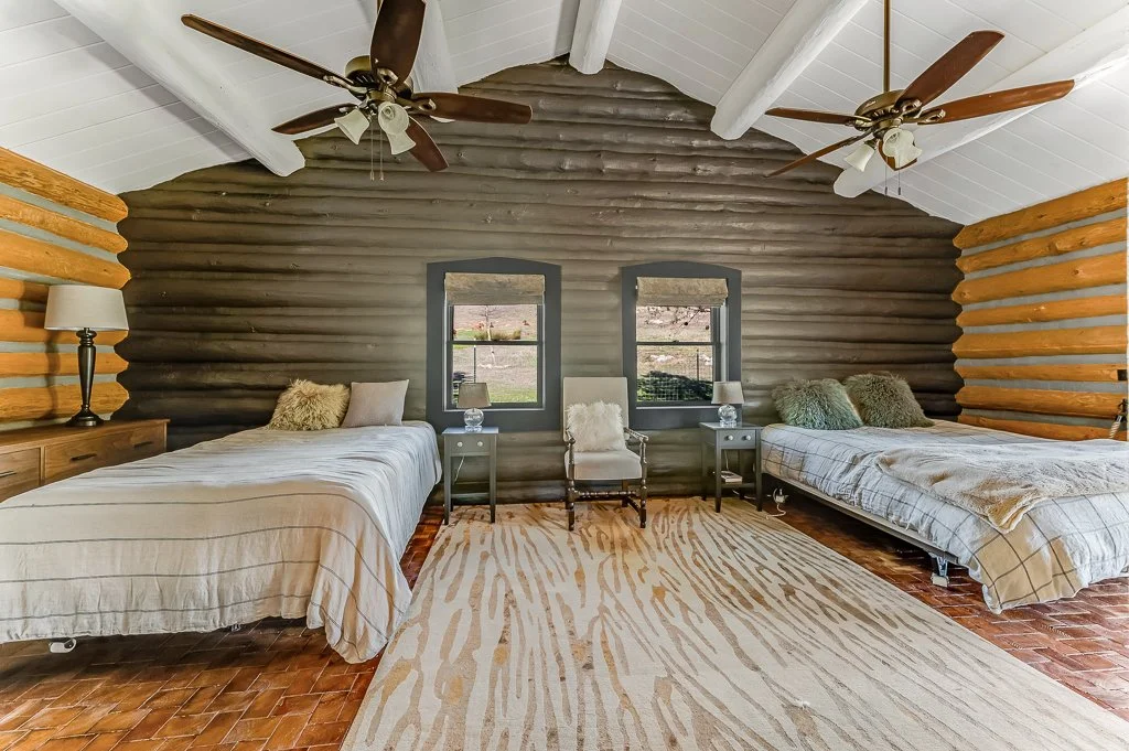 A spacious bedroom with rustic log walls, two beds with bedding and pillows, two windows, two nightstands with lamps, a central armchair, ceiling fans, and a patterned area rug on hardwood flooring.