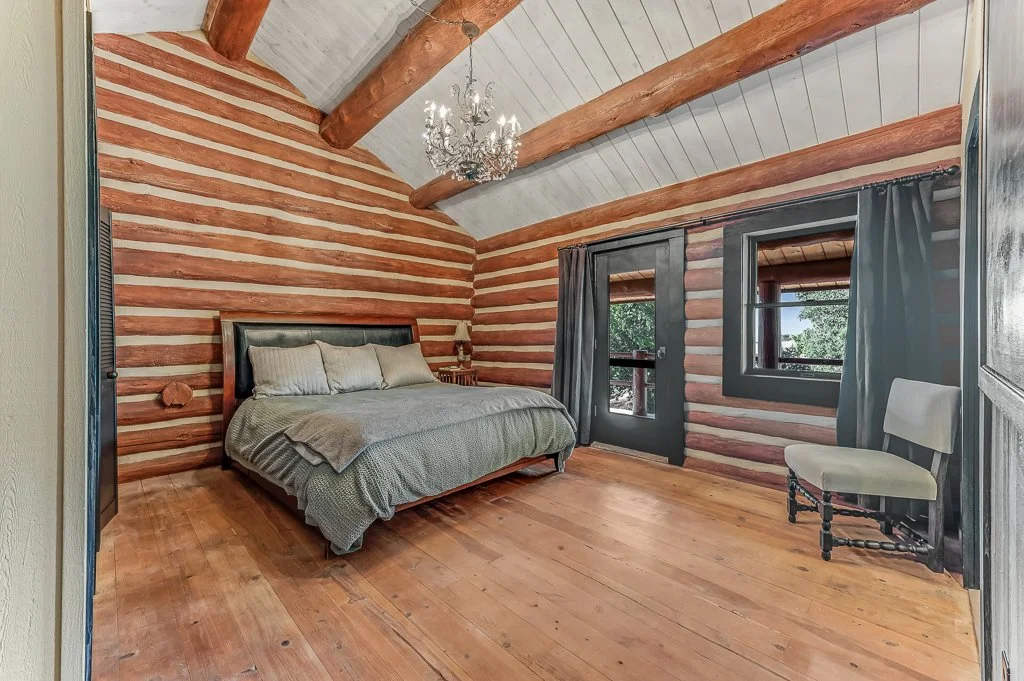 A rustic bedroom with a wood-paneled wall and ceiling, a chandelier, a bed with pillows, a nightstand with a lamp, dark-framed windows with curtains, and a wooden chair.