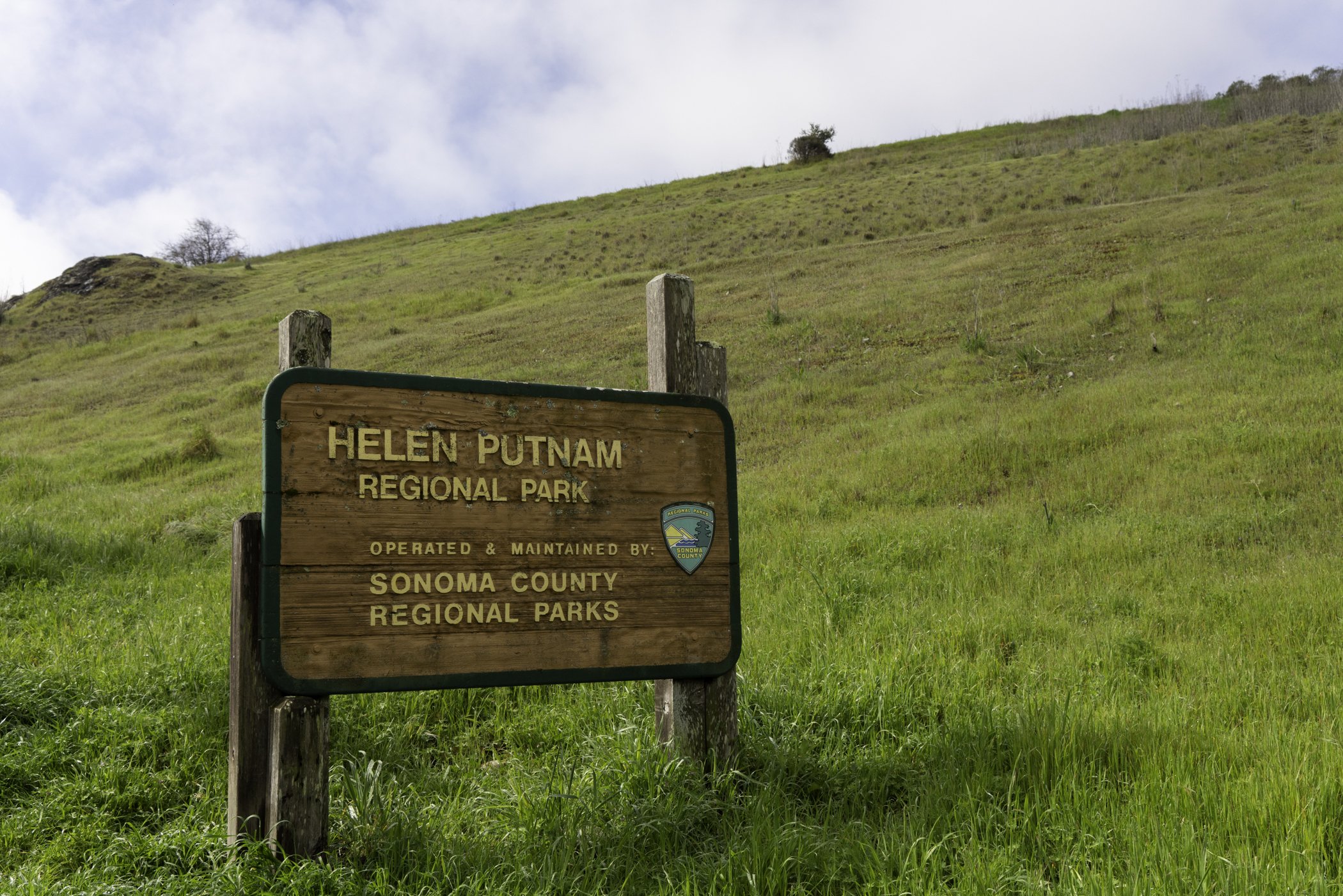 Wooden sign for Helen Putnam Regional Park operated by Sonoma County Regional Parks, set against grassy hillside with scattered small bushes and a partly cloudy sky.