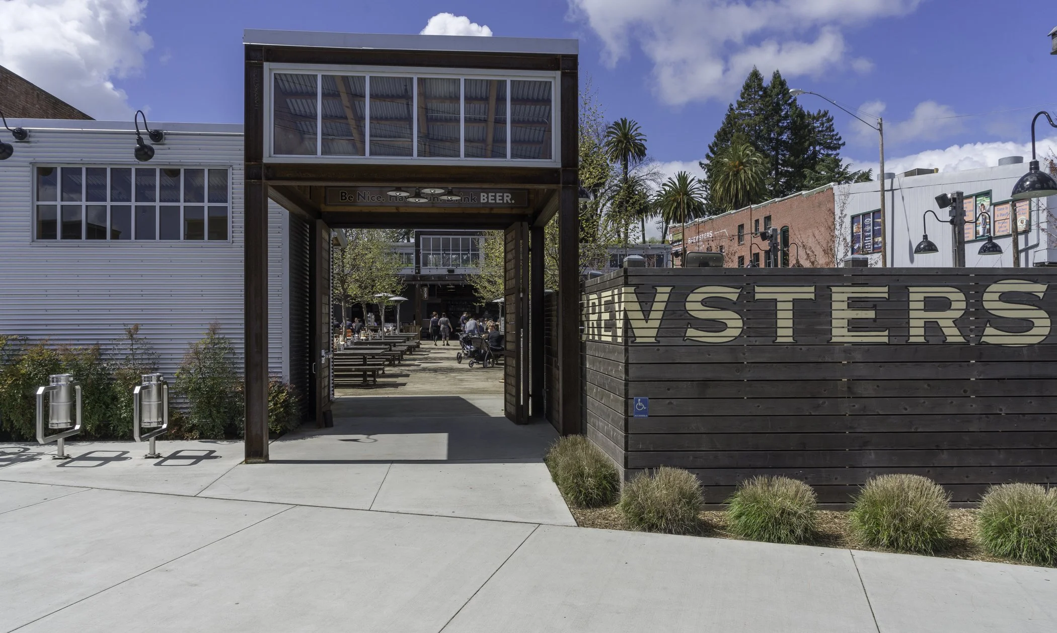 Entrance to a restaurant or bar called Brewsters, with outdoor seating area visible inside. Sign at the entrance promotes being nice and drinking beer. Clear sky and some trees in the background.