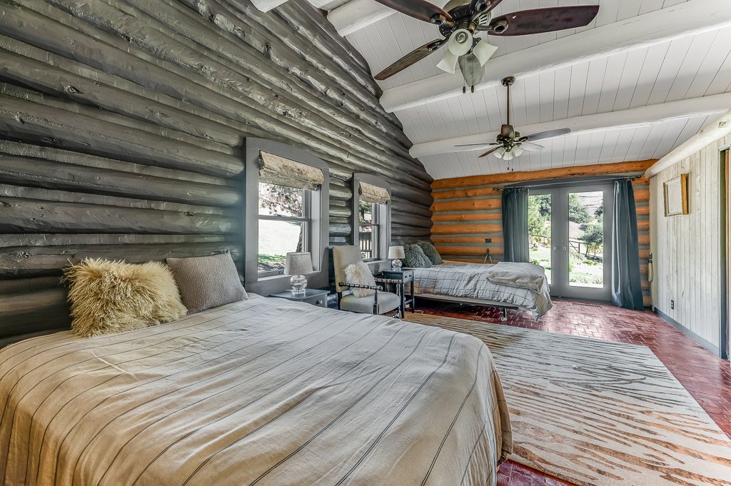 A spacious bedroom with two beds, one on the left with beige bedding and the other on the right with light-colored bedding, a wooden wall, and large windows with curtains.