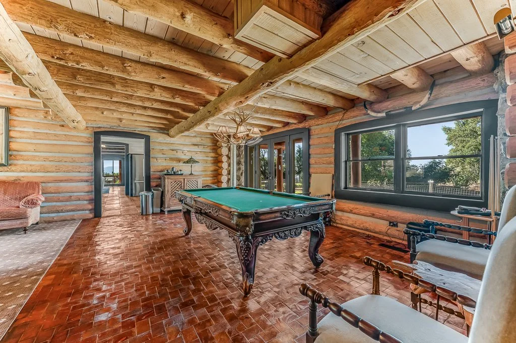 Spacious rustic game room with a pool table, wooden log walls and ceiling, large windows, vintage chairs, and a view of trees outside.