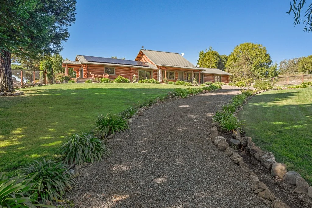 A gravel pathway leading to a log cabin-style house surrounded by green grass and trees under a clear blue sky.