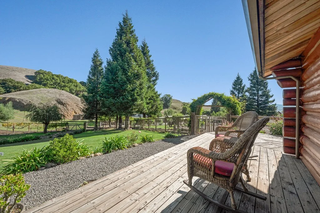 A wooden porch with wicker rocking chairs overlooking a lush green yard with trees and a vine-covered arch.