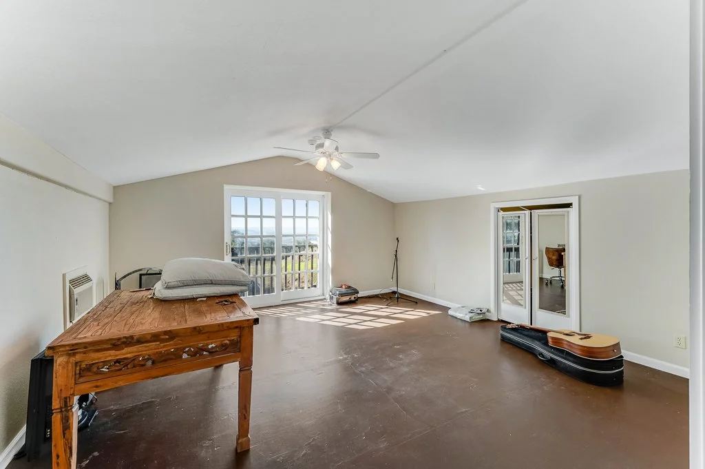 Empty room with a wooden table, a pillow on top, a music stand, guitar case, and a balcony door letting in natural light.