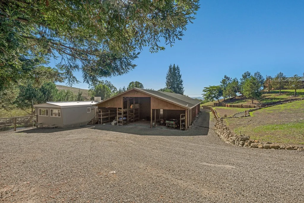 A rural property features a gravel driveway leading to a wooden shed with a metal roof, surrounded by trees and green hills under a clear blue sky.