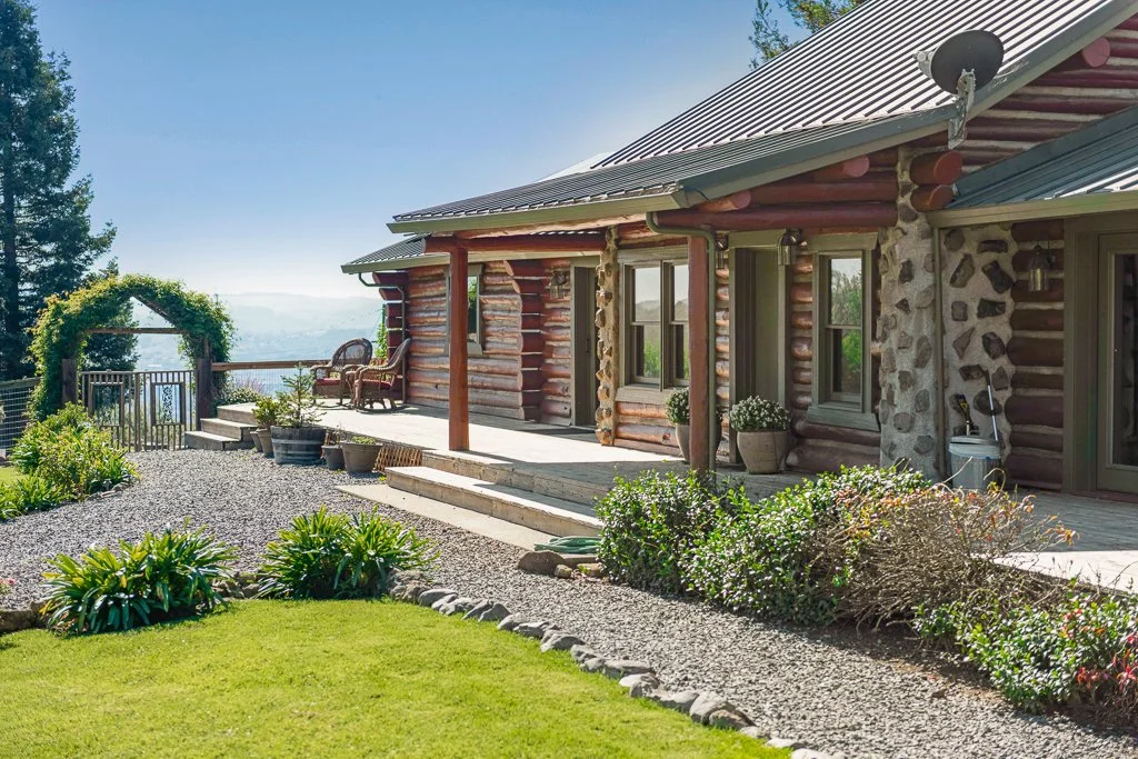 A rustic log cabin with a metal roof sits on a landscaped hillside with a gravel pathway, potted plants, and outdoor seating. An archway covered with greenery leads to a view of the water and trees in the distance.