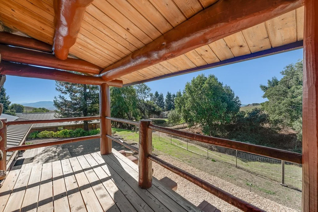 View from a wooden balcony with a rustic log railing, overlooking a backyard with trees, a fence, and distant hills under a clear blue sky.