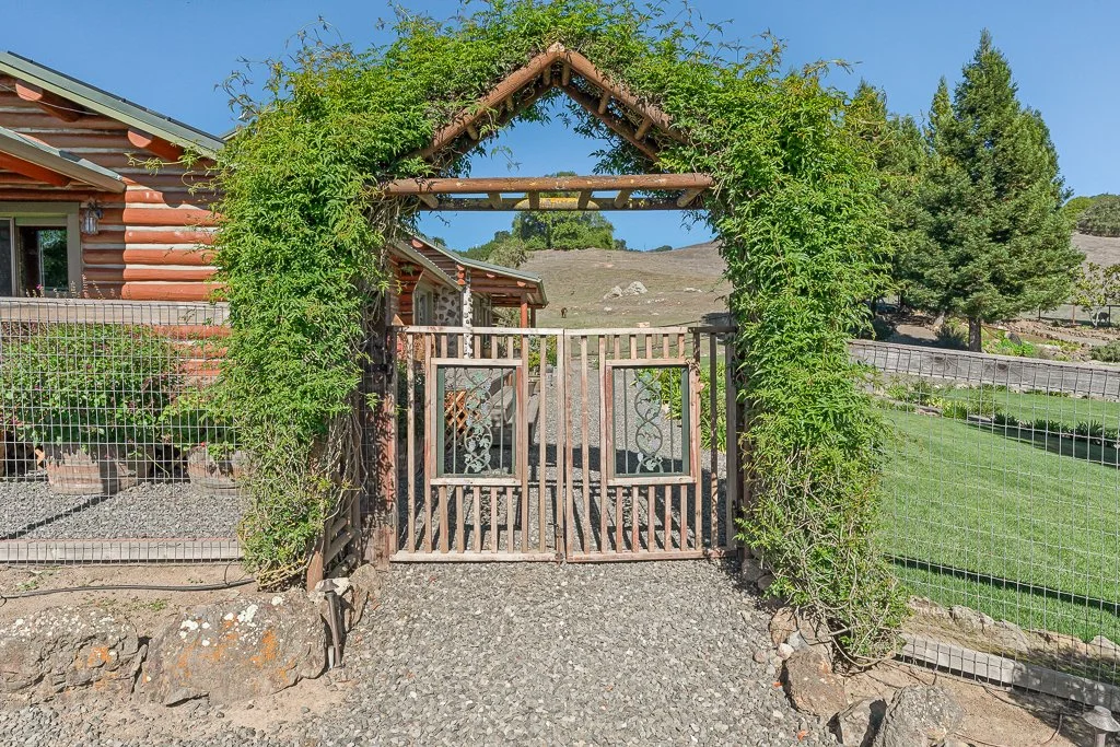 Wooden gate covered with green vines, leading into a yard with a cottage-style house on the left and a large grassy area on the right.