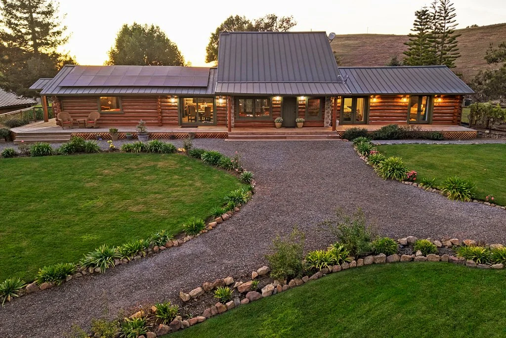 A large wooden house with a metal roof, surrounded by landscaped gardens and a gravel driveway, at sunset.
