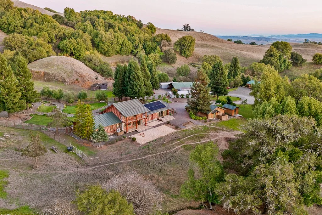 Aerial view of a rural property with a main house featuring solar panels, surrounded by trees, hills, and smaller outbuildings, with a fenced garden area and a driveway.