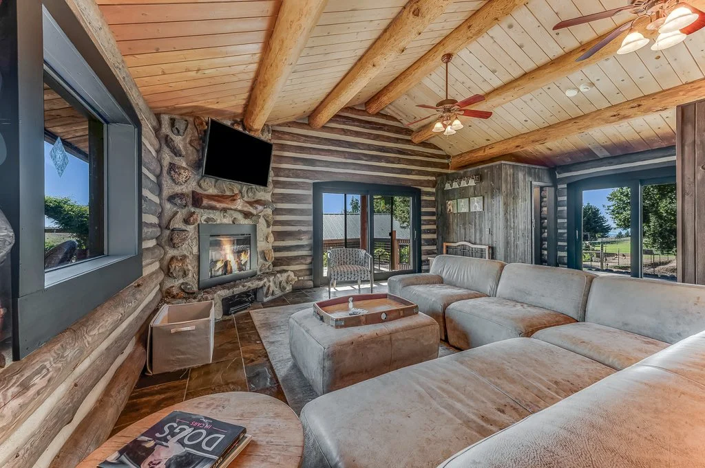 Living room with large sectional sofa, stone fireplace, wooden ceiling with beams, ceiling fans, window, and sliding door leading outside.
