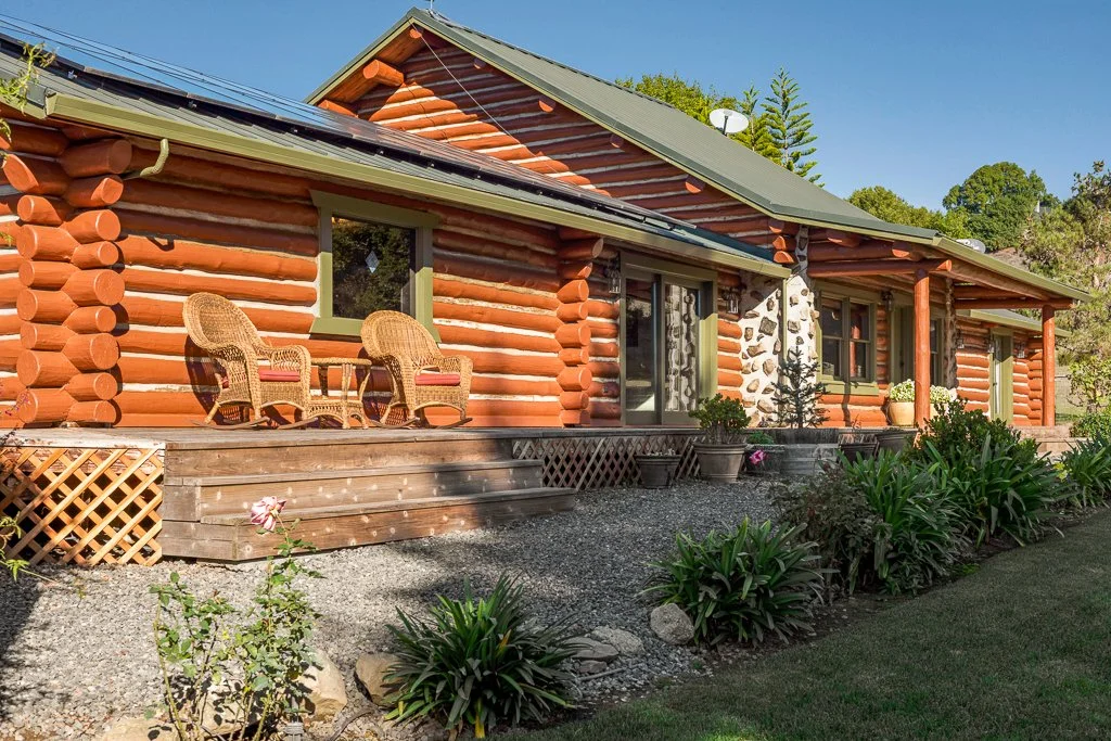 Log cabin with a green metal roof and a small porch with two wicker chairs, surrounded by garden plants and gravel.