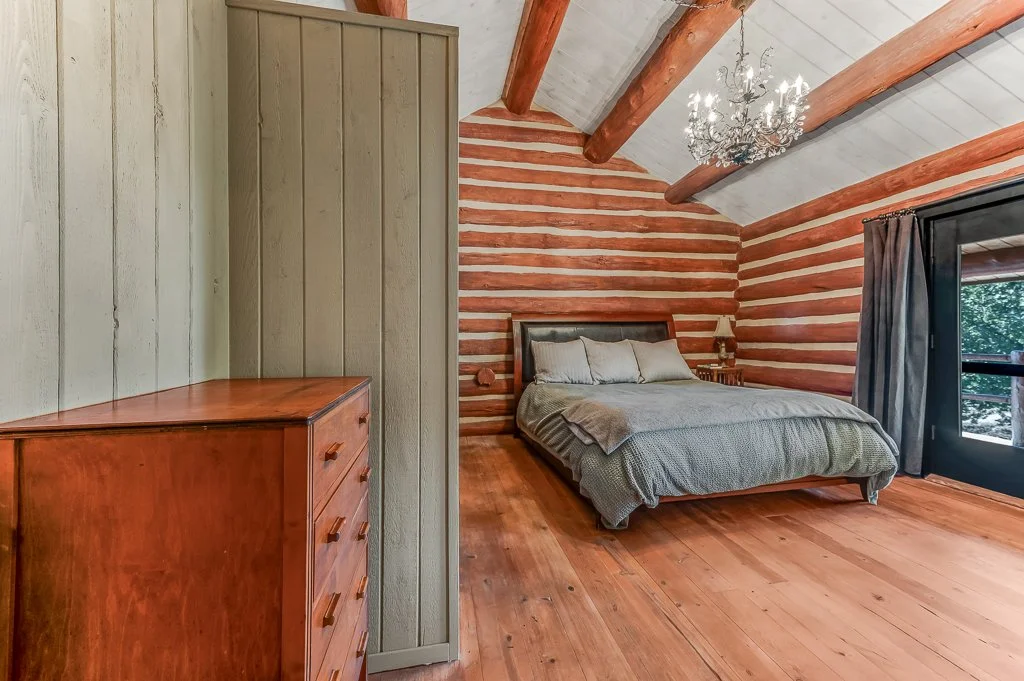 Bedroom with wooden floor and log cabin walls, a large bed with pillows, a small nightstand with a lamp, a window with curtains, and a chandelier hanging from the ceiling.