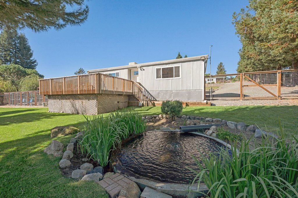 Backyard with a small pond, green grass, a wooden deck attached to a white house, and a fenced yard under a clear blue sky.