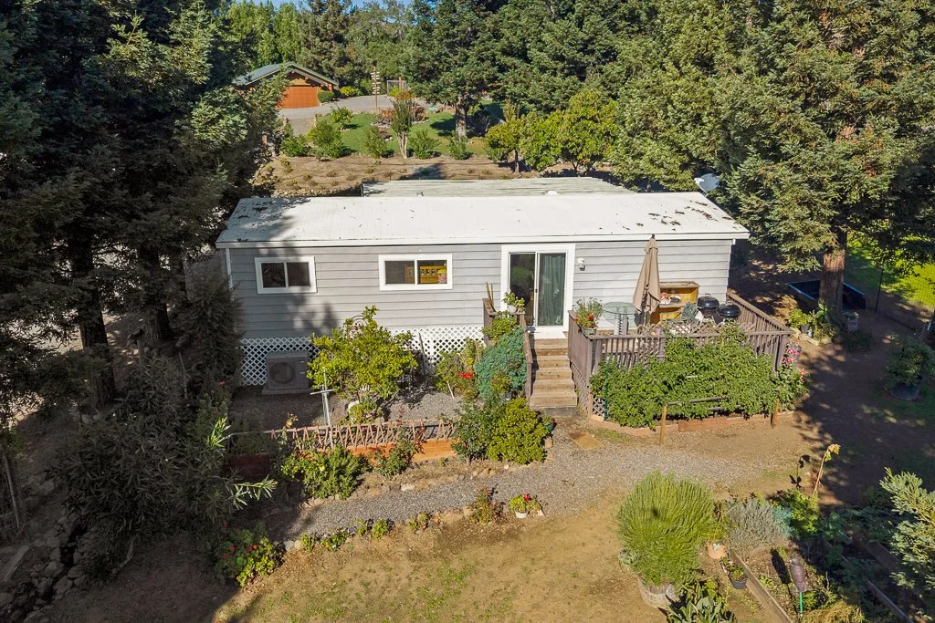 Aerial view of a small house with a white exterior and metal roof, surrounded by trees, shrubs, and garden areas, with a patio and outdoor furniture.