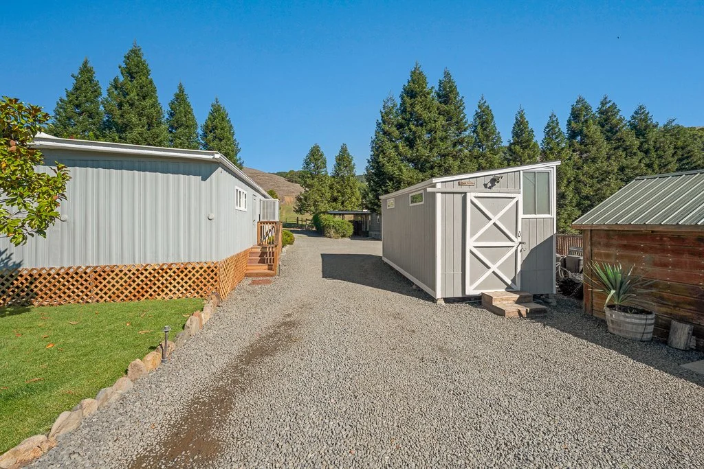 A gravel driveway flanked by a white shed with a wooden deck on the left and a smaller gray shed on the right, with trees and blue sky in the background.