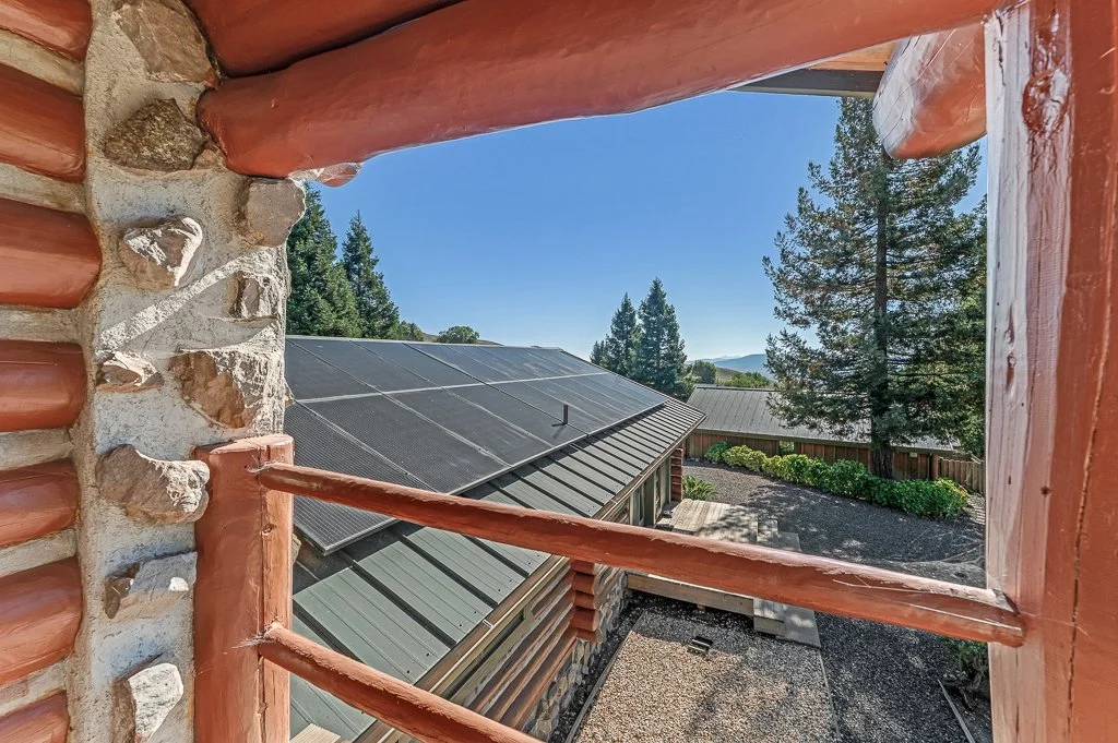 View through a rustic window opening showing a building with solar panels on the roof, surrounded by trees and a clear blue sky.