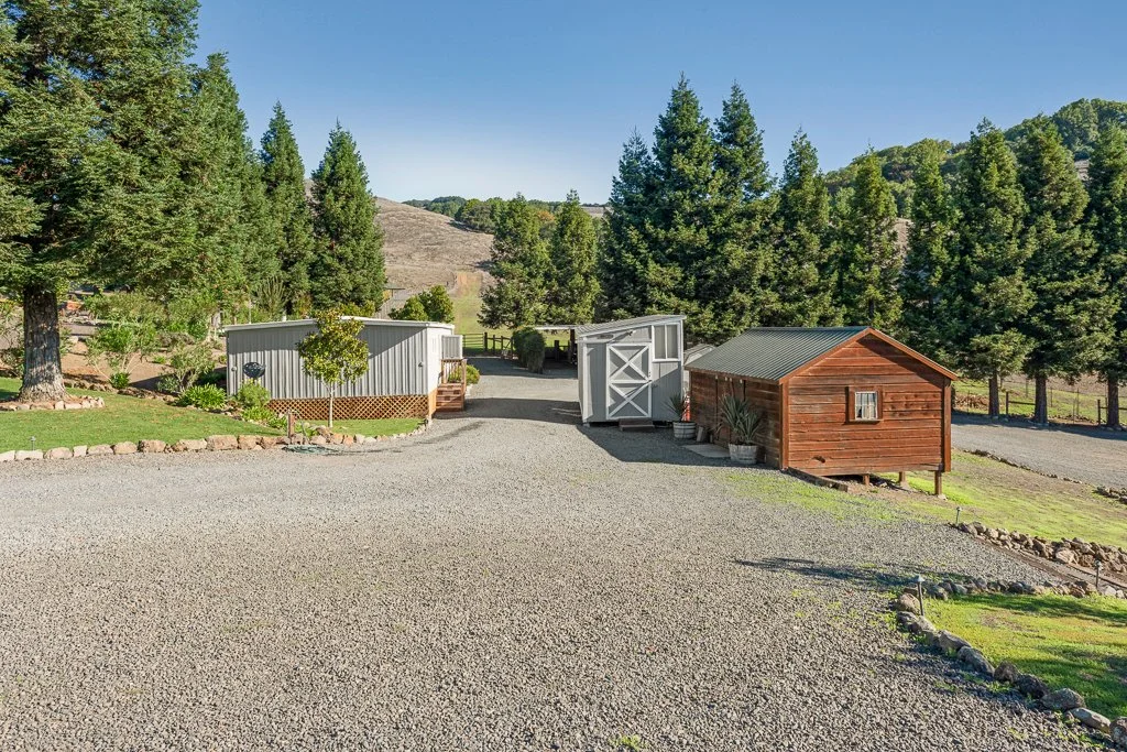 A gravel driveway leading to a white shed with double doors and a small wooden shed, surrounded by trees and greenery under a clear blue sky.