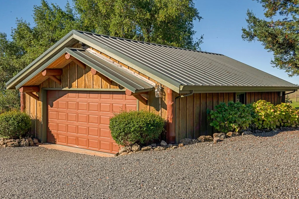 A rustic garage with a brown roll-up door, wooden siding, and a metal roof surrounded by greenery and gravel.