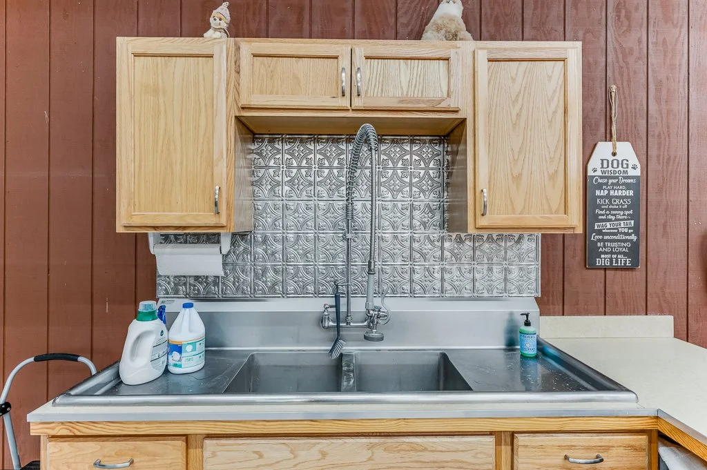 Kitchen sink with cleaning supplies, soap dispenser, wooden cabinets, a decorative wall plaque, and a tiled backsplash.