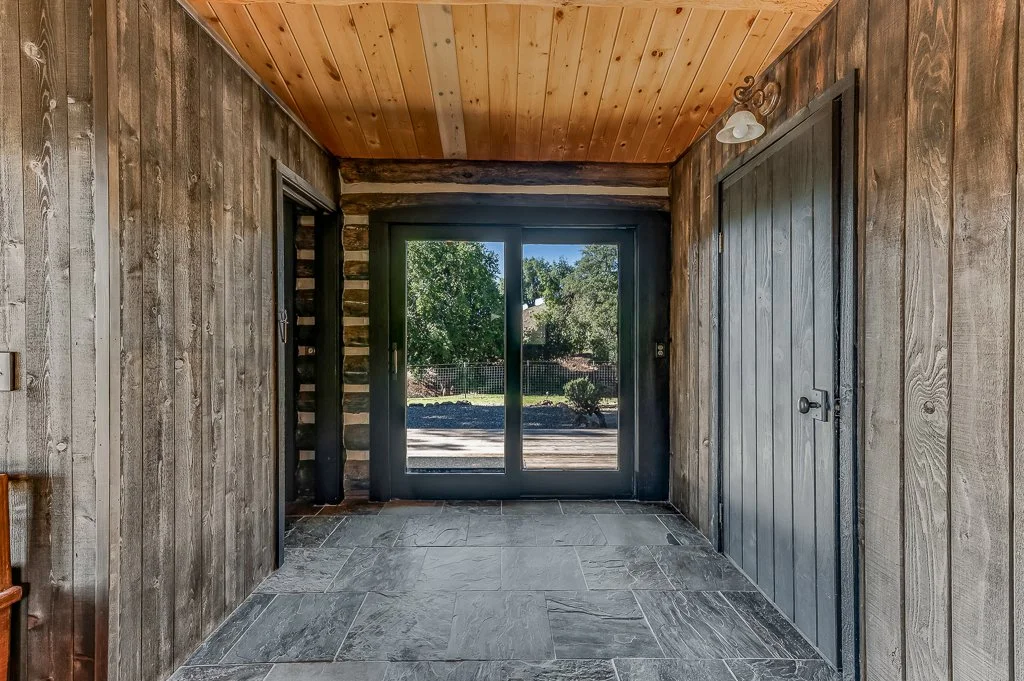 Interior view of a rustic room with wood-paneled walls and ceiling, stone tile flooring, and a central sliding glass door leading outside to a yard with trees.
