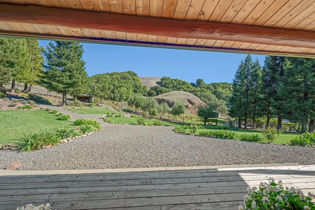 View of a landscaped outdoor yard with green grass, trees, and bushes, seen from a wooden porch, with hills and a clear blue sky in the background.