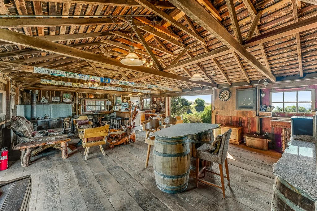 Interior of a rustic bar or lounge area with wooden furniture, a barrel table, a dartboard, and large windows showing a scenic view outside.