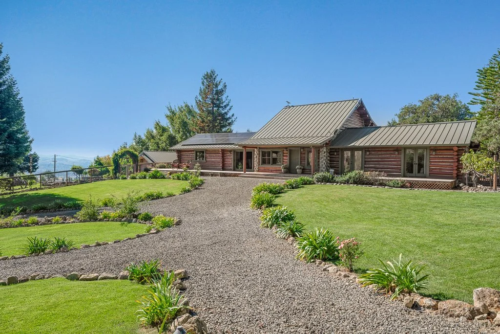 A scenic view of a ranch-style house with a gravel driveway, surrounded by green lawns and landscaped plants under a clear blue sky.