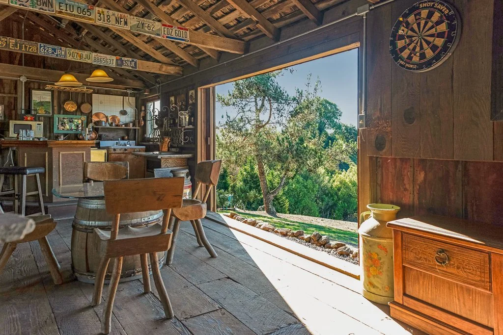 Interior of a rustic wooden cabin with a view of a lush outdoor landscape, featuring a dartboard on the wall, a small bar area, wooden chairs, and vintage decor.